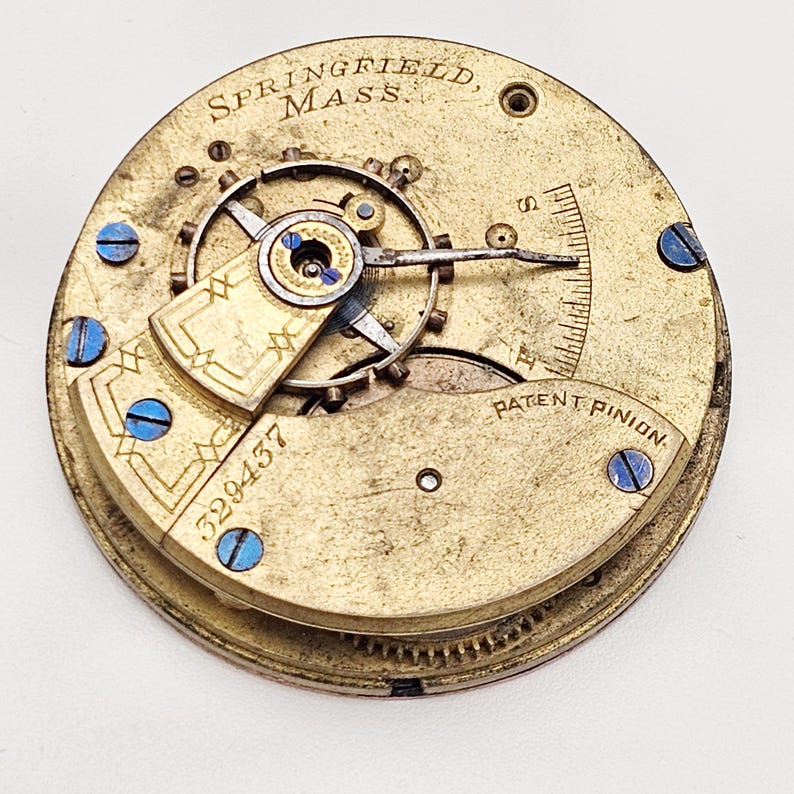 Puede incluir: Primer plano de un movimiento de reloj antiguo. El mecanismo de color lat&oacute;n presenta engranajes intrincados, un volante y tornillos azules. El texto "SPRINGFIELD, MASS." y "PATENT PINION" son visibles. El n&uacute;mero de serie "329437" tambi&eacute;n est&aacute; presente.