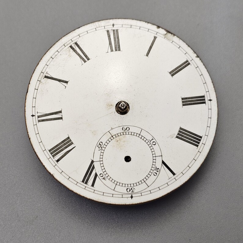 Puede incluir: Esfera de reloj de bolsillo blanca con marcadores de hora en n&uacute;meros romanos y marcas negras. La esfera tiene un c&iacute;rculo m&aacute;s peque&ntilde;o en el centro con marcas negras para los segundos.
