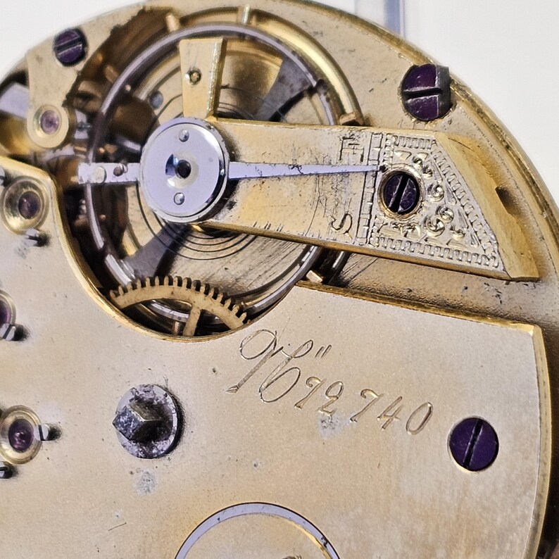 Może przedstawiać: Zbliżenie na mechanizm kieszonkowego zegarka w kolorze złotym z misternymi kołami zębatymi i dźwignią. Zegarek jest oznaczony numerem "7012740".
