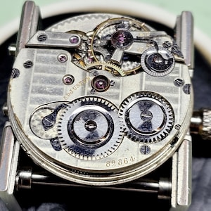 Puede incluir: Primer plano de un movimiento de reloj plateado con el texto "ADJUSTED" y el n&uacute;mero "89864" visible. El movimiento es intrincado y detallado, con muchos engranajes y resortes.