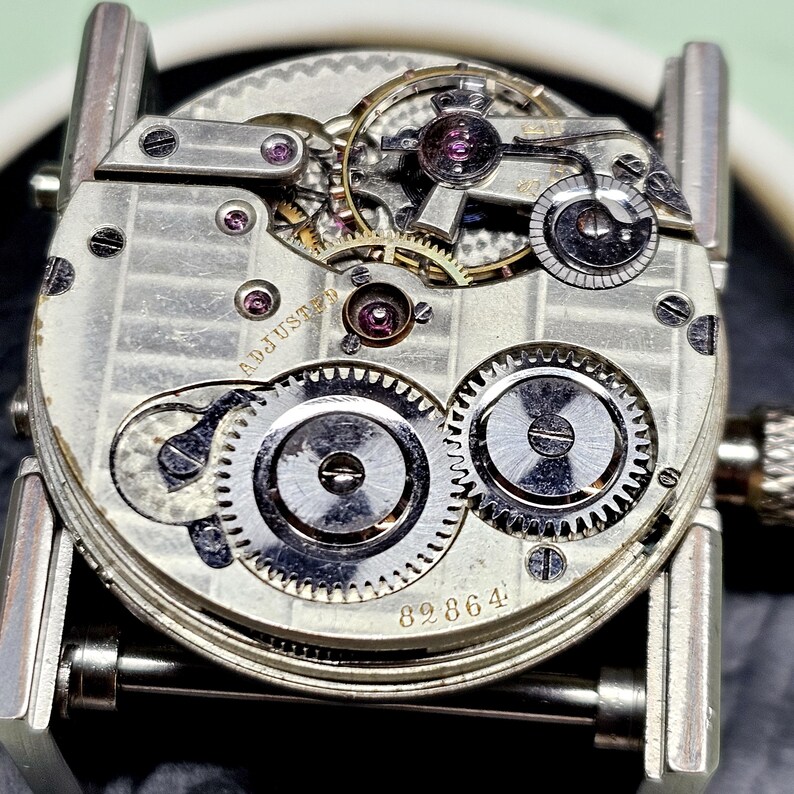 Puede incluir: Primer plano de un movimiento de reloj plateado con el texto "ADJUSTED" y el n&uacute;mero "89864" visible. El movimiento es intrincado y detallado, con muchos engranajes y resortes.