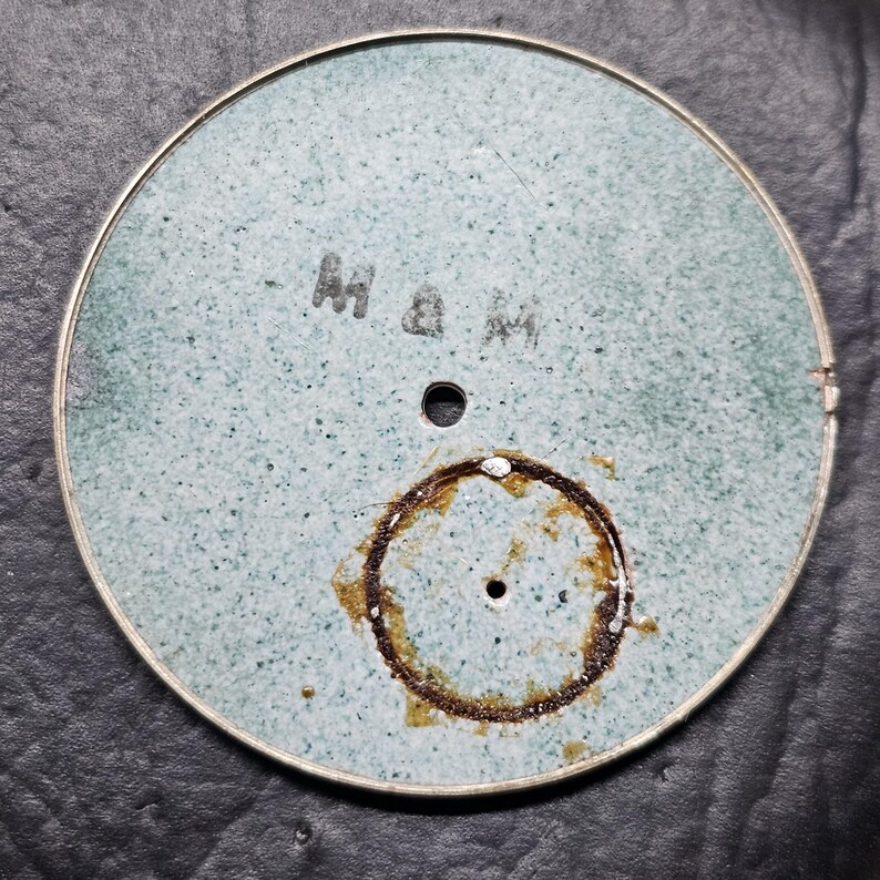 Puede incluir: Una esfera de reloj redonda de color azul claro con un anillo marr&oacute;n alrededor del agujero central. La esfera del reloj tiene el texto "M & M" escrito en ella.
