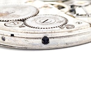 May include: Close-up of a vintage watch movement, showcasing intricate gears and components. The silver-toned metal parts are detailed, with a small, dark jewel set into the frame. The image highlights the precision and craftsmanship of the watch mechanism.