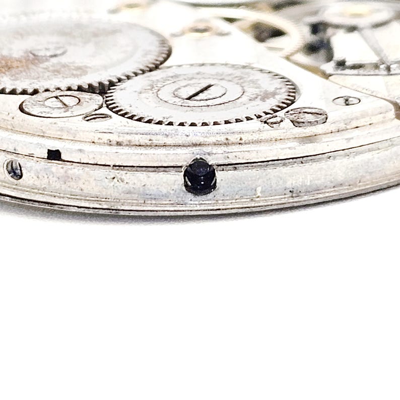 May include: Close-up of a vintage watch movement, showcasing intricate gears and components. The silver-toned metal parts are detailed, with a small, dark jewel set into the frame. The image highlights the precision and craftsmanship of the watch mechanism.