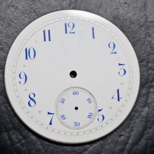 Puede incluir: Esfera de reloj blanca con marcadores de hora en n&uacute;meros romanos azules y marcadores de minutos dorados. La esfera del reloj tiene un c&iacute;rculo conc&eacute;ntrico m&aacute;s peque&ntilde;o con n&uacute;meros negros para los segundos.