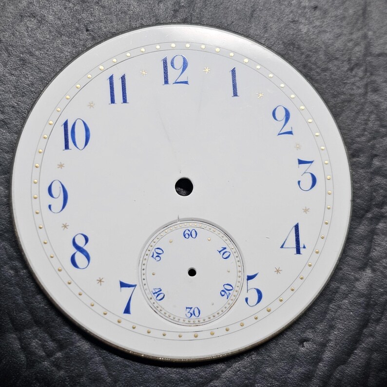 Puede incluir: Esfera de reloj blanca con marcadores de hora en n&uacute;meros romanos azules y marcadores de minutos dorados. La esfera del reloj tiene un c&iacute;rculo conc&eacute;ntrico m&aacute;s peque&ntilde;o con n&uacute;meros negros para los segundos.