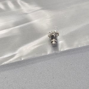 May include: A small silver-colored watch part with a gear mechanism and a round shaft.