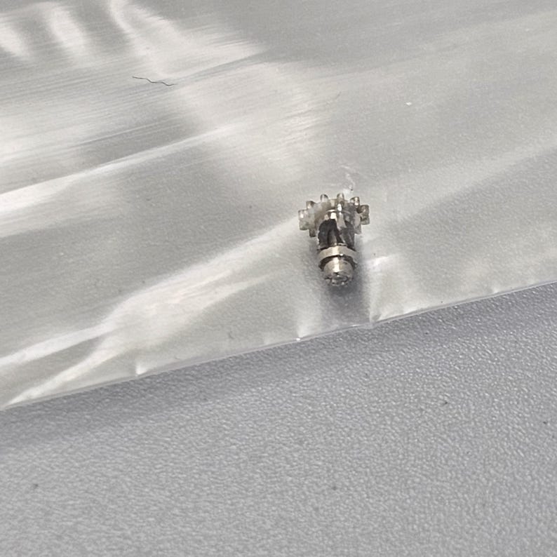 May include: A small silver-colored watch part with a gear mechanism and a round shaft.