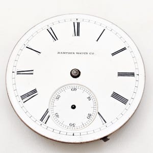 Puede incluir: Una esfera de reloj de bolsillo blanca con n&uacute;meros romanos negros y un peque&ntilde;o dial de segundos. Las palabras "HAMPDEN WATCH CO" est&aacute;n impresas en negro. La esfera tiene un borde met&aacute;lico y un peque&ntilde;o accesorio en la parte inferior.