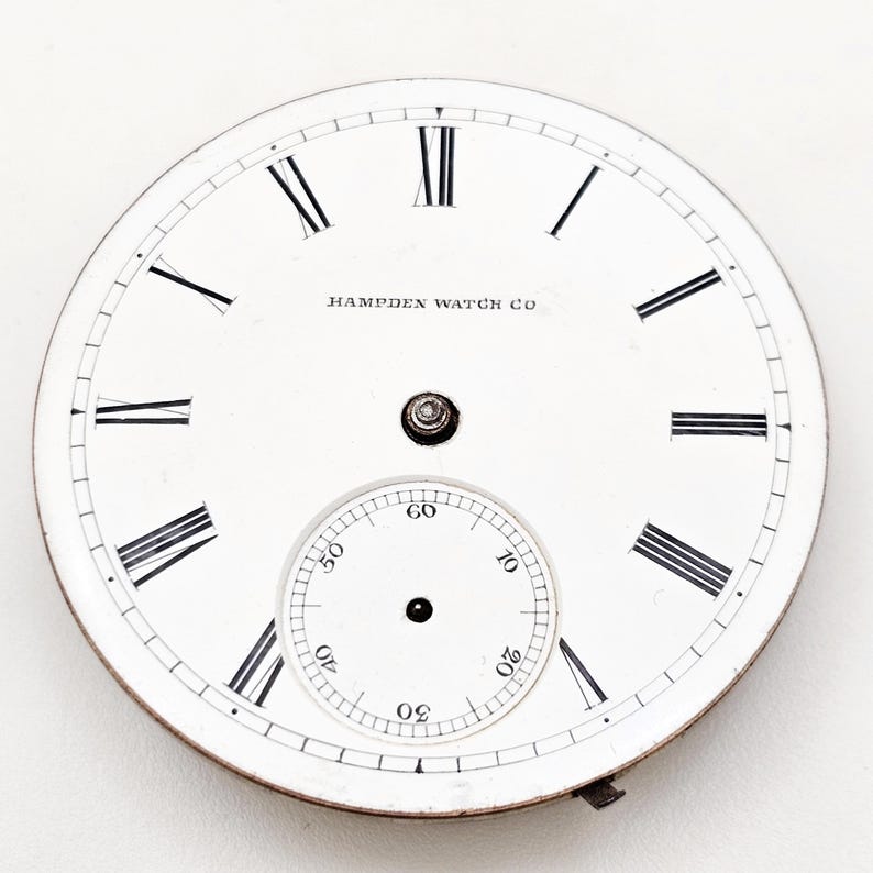 Puede incluir: Una esfera de reloj de bolsillo blanca con n&uacute;meros romanos negros y un peque&ntilde;o dial de segundos. Las palabras "HAMPDEN WATCH CO" est&aacute;n impresas en negro. La esfera tiene un borde met&aacute;lico y un peque&ntilde;o accesorio en la parte inferior.