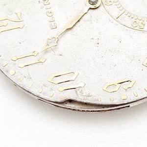 May include: Close-up of a vintage Elgin watch face. The silver-toned face features gold-colored numerals and hands. The word "ELGIN" is visible at the top. The watch face shows signs of age and wear.