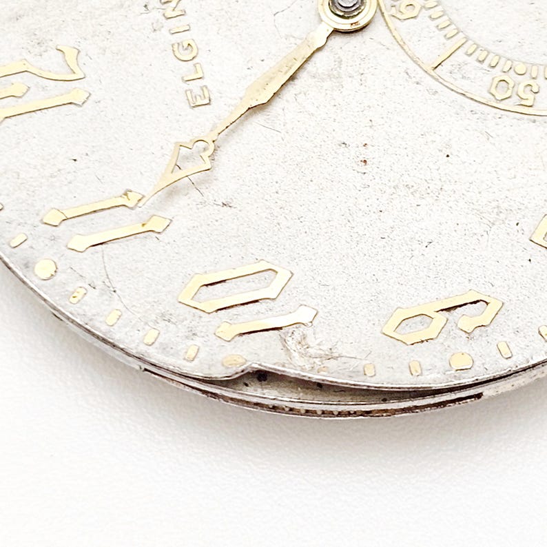 May include: Close-up of a vintage Elgin watch face. The silver-toned face features gold-colored numerals and hands. The word "ELGIN" is visible at the top. The watch face shows signs of age and wear.