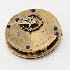 May include: A vintage watch movement with intricate gears and components. The brass-colored mechanism features blue screws and engraved text, including "North Watch Co." and the serial number 1404669. The watch movement is set against a white background.