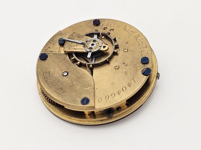 May include: A vintage watch movement with intricate gears and components. The brass-colored mechanism features blue screws and engraved text, including "North Watch Co." and the serial number 1404669. The watch movement is set against a white background.