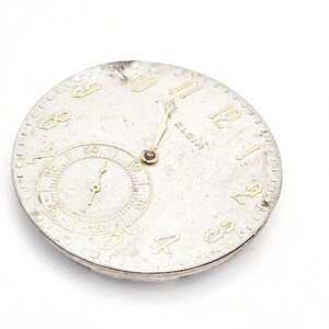 May include: A vintage Elgin watch face with a silver-colored dial and gold-toned numerals and hands. A smaller subdial is at the bottom. The watch face has a slightly aged appearance.