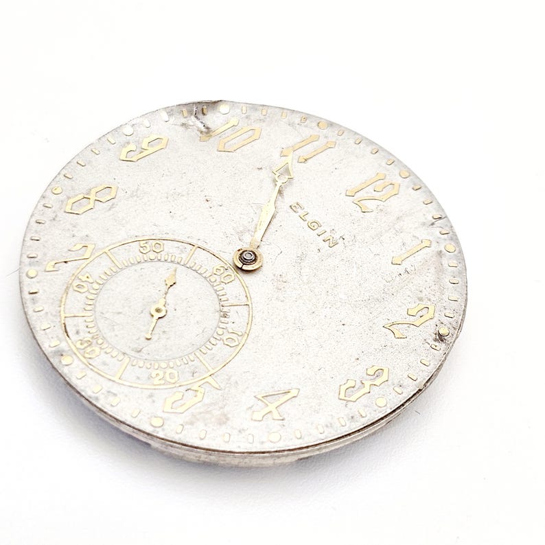 May include: A vintage Elgin watch face with a silver-colored dial and gold-toned numerals and hands. A smaller subdial is at the bottom. The watch face has a slightly aged appearance.