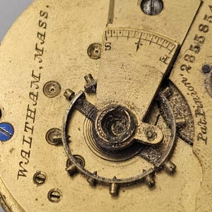 Puede incluir: Primer plano de un movimiento de reloj de color dorado con la inscripci&oacute;n "WALTHAM, MASS." y un peque&ntilde;o indicador circular etiquetado como "S" y "E".