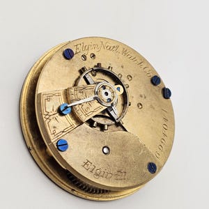 May include: Close-up of a vintage Elgin Natl. Watch Co. pocket watch movement. The gold-toned mechanism features intricate gears, blue screws, and the Elgin name. The serial number 1404669 is visible.