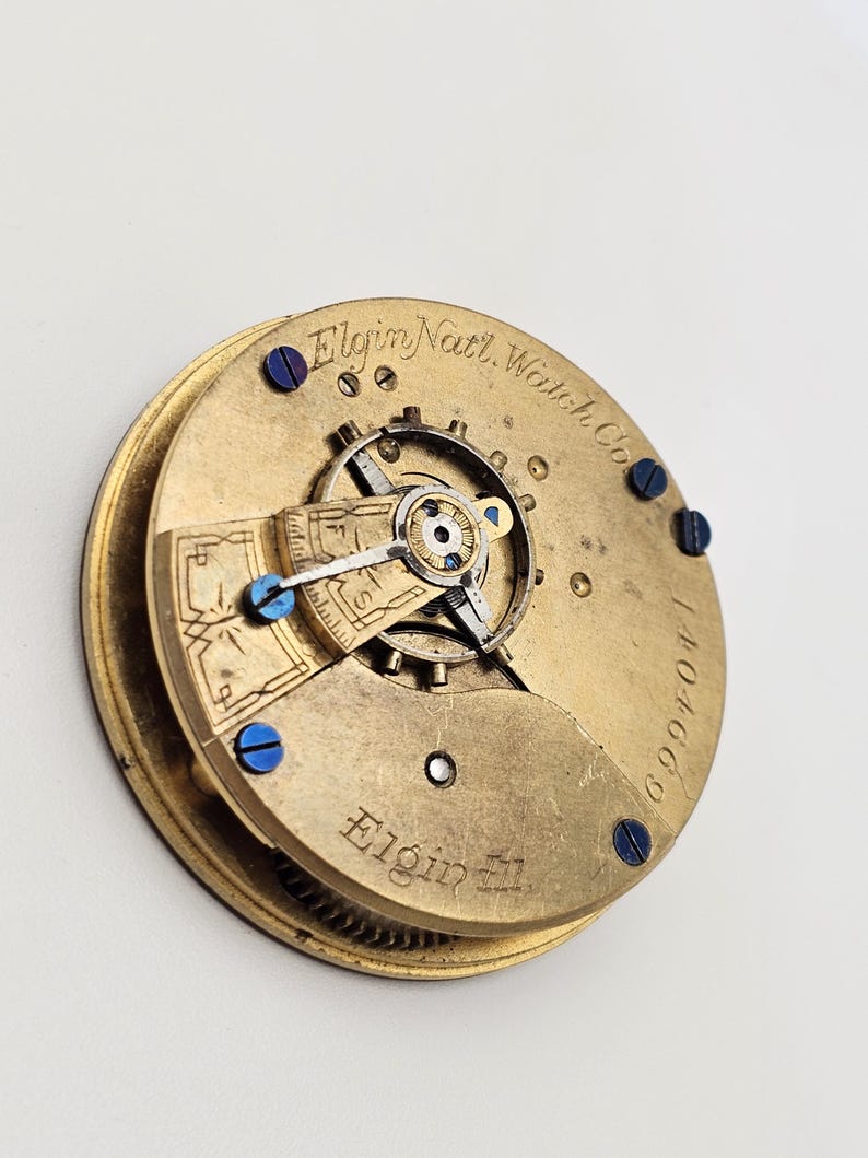 May include: Close-up of a vintage Elgin Natl. Watch Co. pocket watch movement. The gold-toned mechanism features intricate gears, blue screws, and the Elgin name. The serial number 1404669 is visible.