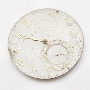 May include: A vintage Elgin watch face with a silver finish and gold-colored numerals and hands. The face includes a smaller subdial and the brand name "Elgin". The watch face is round and shows signs of age.