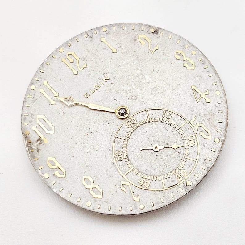 May include: A vintage Elgin watch face with a silver finish and gold-colored numerals and hands. The face includes a smaller subdial and the brand name "Elgin". The watch face is round and shows signs of age.