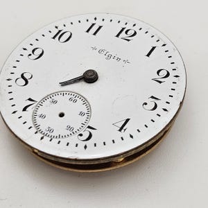 May include: A vintage Elgin pocket watch face with a white background, black numerals, and a smaller seconds dial. The watch face has a gold-colored rim and the word "Elgin" is printed on the face.