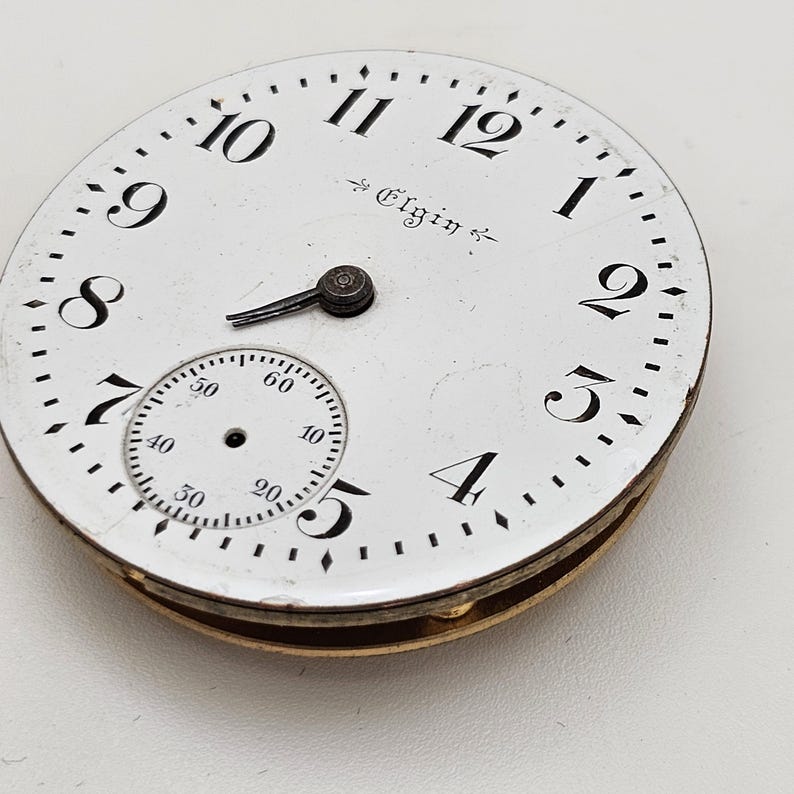 May include: A vintage Elgin pocket watch face with a white background, black numerals, and a smaller seconds dial. The watch face has a gold-colored rim and the word "Elgin" is printed on the face.