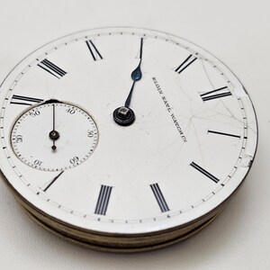 May include: Close-up of an antique Elgin Natl. Watch Co. pocket watch face. The white face features black Roman numerals, a smaller seconds dial, and blue hands. The watch face is cracked and shows signs of age.