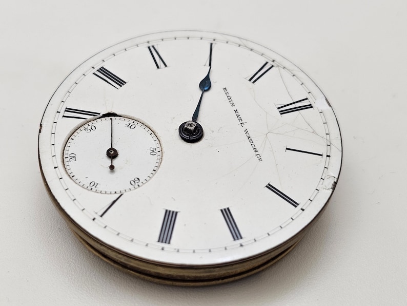 May include: Close-up of an antique Elgin Natl. Watch Co. pocket watch face. The white face features black Roman numerals, a smaller seconds dial, and blue hands. The watch face is cracked and shows signs of age.