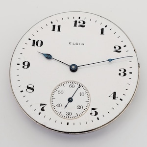 以下が含まれることがあります： 黒い数字と「ELGIN」のブランド名が付いた白い時計の文字盤。小さなサブダイヤルと青い針が付いています。時計の文字盤は金属製の縁があり、無地の背景に配置されています。