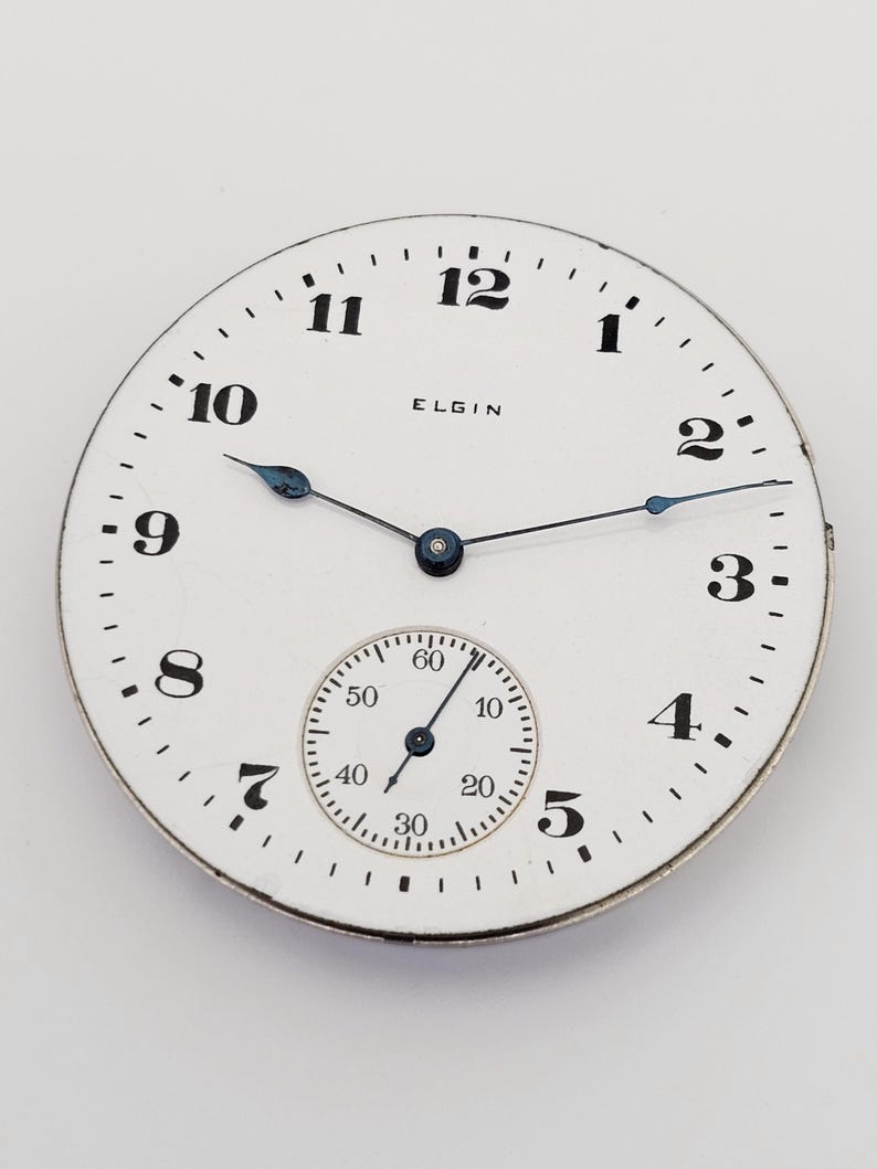 以下が含まれることがあります： 黒い数字と「ELGIN」のブランド名が付いた白い時計の文字盤。小さなサブダイヤルと青い針が付いています。時計の文字盤は金属製の縁があり、無地の背景に配置されています。