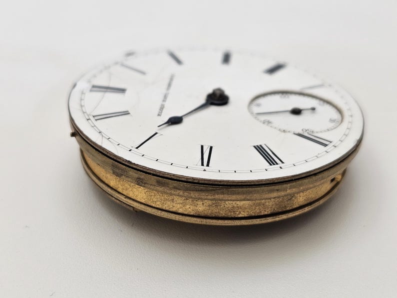 May include: A vintage pocket watch face with a white dial, black Roman numerals, and a smaller subdial. The watch has black hands and a gold-toned bezel. The text "WALTHAM U.S.A. CO." is visible on the dial.