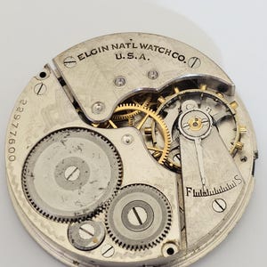 以下が含まれることがあります： Elgin Natl Watch Co. U.S.A. の時計ムーブメントのクローズアップ。銀色のメカニズムは、複数の歯車、ネジ、テンプを備えています。シリアル番号22977600が表示されています。時計のムーブメントは円形です。