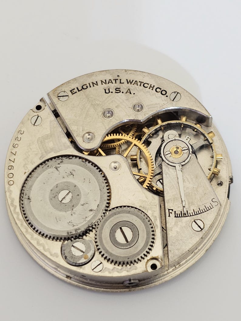 以下が含まれることがあります： Elgin Natl Watch Co. U.S.A. の時計ムーブメントのクローズアップ。銀色のメカニズムは、複数の歯車、ネジ、テンプを備えています。シリアル番号22977600が表示されています。時計のムーブメントは円形です。