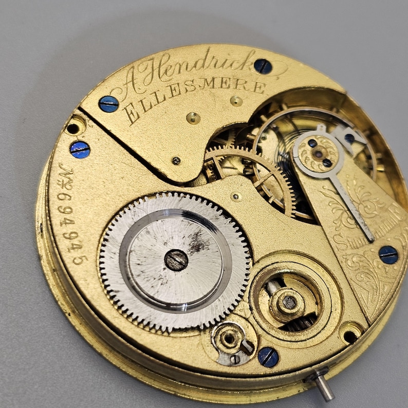 May include: Close-up of a gold-colored pocket watch movement with the inscription "A Hendrick Ellesmere" and the number "694945".