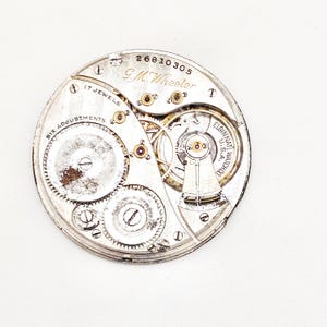 May include: A vintage watch movement with visible gears, jewels, and the text "G.M. Wheeler" and "Elgin Nat'l Watch Co. U.S.A.". The silver-toned mechanism features intricate details, including "17 Jewels" and "Six Adjustments".