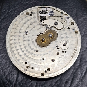 Puede incluir: Primer plano de la parte posterior de un reloj de bolsillo plateado con un sistema de engranajes intrincado y el n&uacute;mero 028064 impreso en la parte posterior.