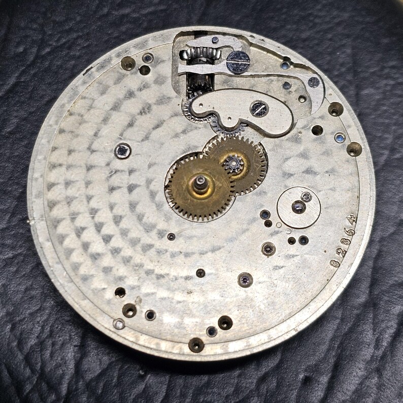 Puede incluir: Primer plano de la parte posterior de un reloj de bolsillo plateado con un sistema de engranajes intrincado y el n&uacute;mero 028064 impreso en la parte posterior.