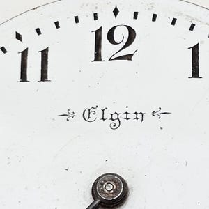 May include: Close-up of a white clock face with black numerals and markings. The number 12 is prominently displayed at the top, with the word "Elgin" in elegant script below. A small, circular component is at the bottom.