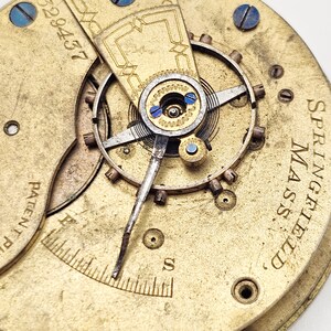 Puede incluir: Primer plano de un mecanismo de reloj antiguo, con engranajes dorados y detalles intrincados. La esfera del reloj incluye el texto "SPRINGFIELD MASS." y el n&uacute;mero "29457". El dise&ntilde;o presenta tornillos azules y un peque&ntilde;o dial con marcas.