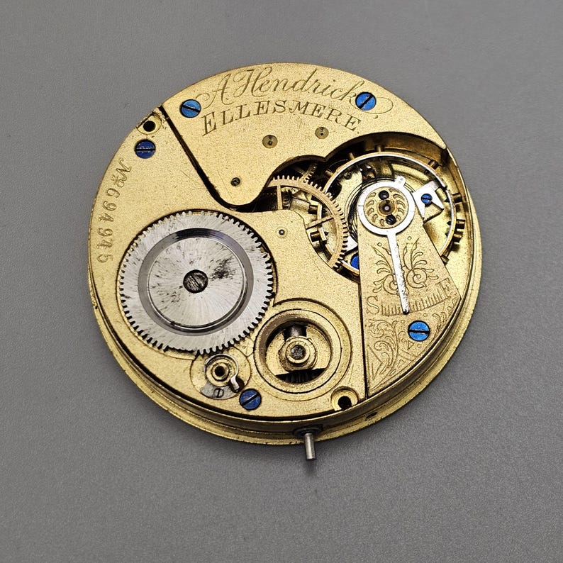 May include: A close-up view of the inner workings of a gold pocket watch. The watch face is engraved with the text "A Hendrick Ellesmere" and the number "542694". The watch has blue screws and a decorative floral design.