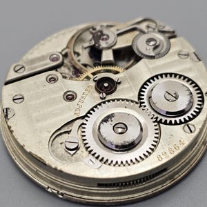 Puede incluir: Primer plano de un movimiento de reloj de bolsillo plateado con engranajes intrincados y la inscripci&oacute;n "ADJUSTED" visible. La esfera del reloj est&aacute; marcada con el n&uacute;mero "89864".