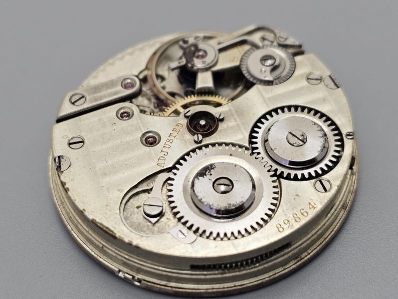 Puede incluir: Primer plano de un movimiento de reloj de bolsillo plateado con engranajes intrincados y la inscripci&oacute;n "ADJUSTED" visible. La esfera del reloj est&aacute; marcada con el n&uacute;mero "89864".