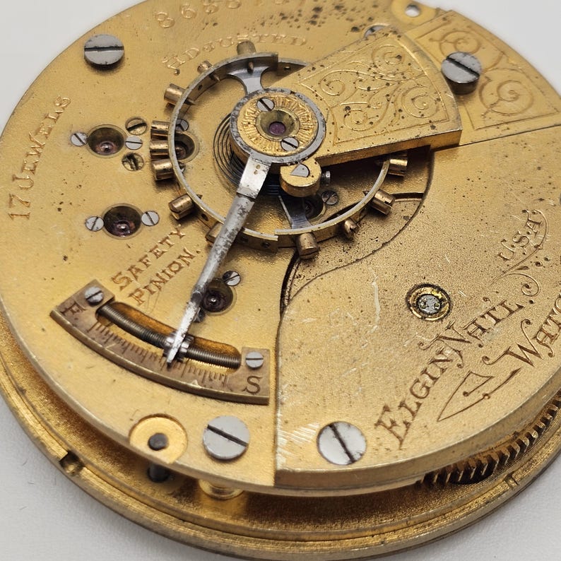 May include: Close-up of a vintage Elgin National Watch Company watch movement. The gold-toned mechanism features intricate gears, screws, and a balance wheel. Text includes "17 Jewels" and "Safety Pinion". The watch movement is a detailed example of early 20th-century watchmaking.