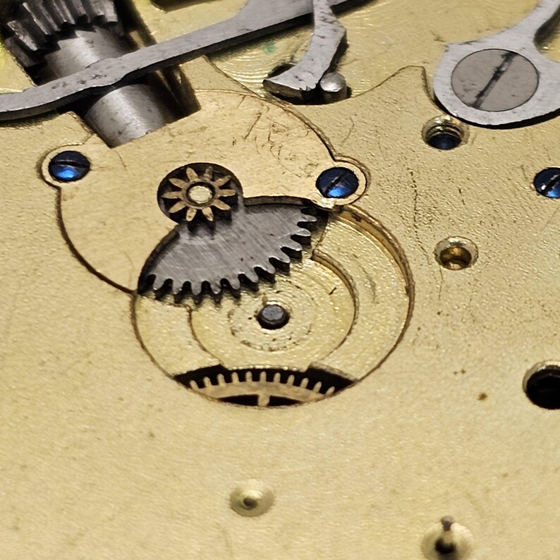 May include: Close-up of a gold-colored watch mechanism with a silver gear and a smaller star-shaped gear. The mechanism is surrounded by a circular, concentric pattern of rings.