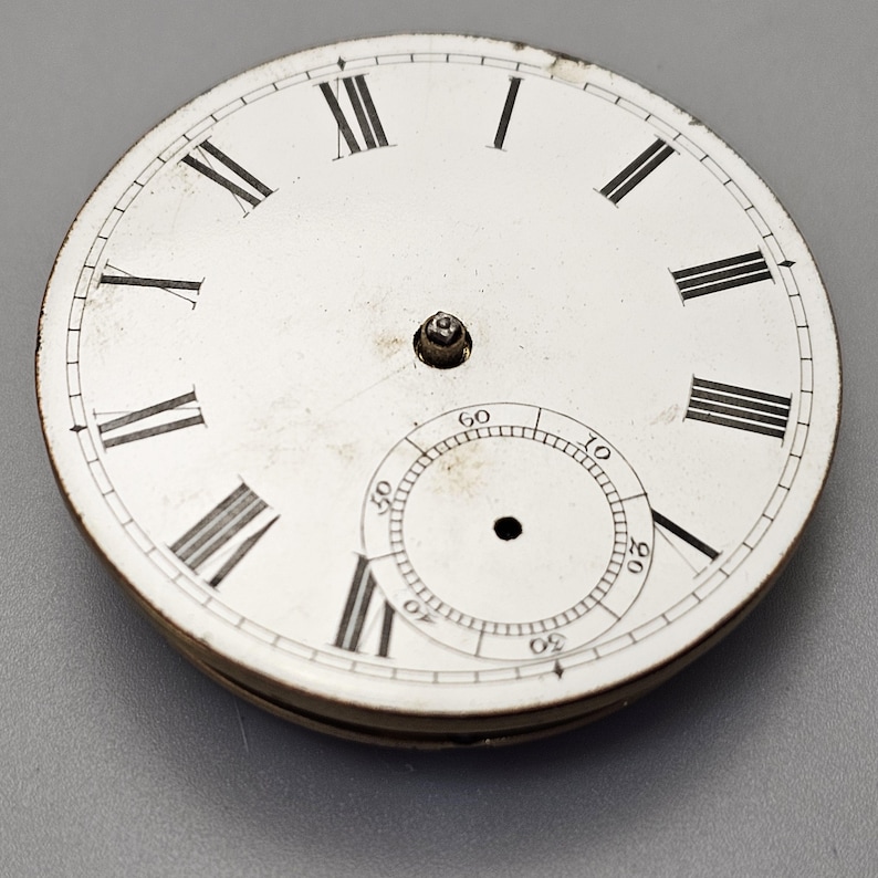 Puede incluir: Una esfera de reloj de bolsillo blanca con marcadores de hora en n&uacute;meros romanos y un c&iacute;rculo conc&eacute;ntrico m&aacute;s peque&ntilde;o con marcas de minutos. La esfera del reloj est&aacute; ligeramente sucia y tiene un peque&ntilde;o punto negro en el centro.