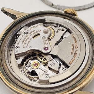 May include: Close-up of a vintage Gruen watch movement. The watch mechanism is visible, showcasing intricate gears, jewels, and the "AUTOWIND VERI-THIN" inscription. The watch case is gold-toned, and the movement is marked "SEVENTEEN 17 JEWELS".