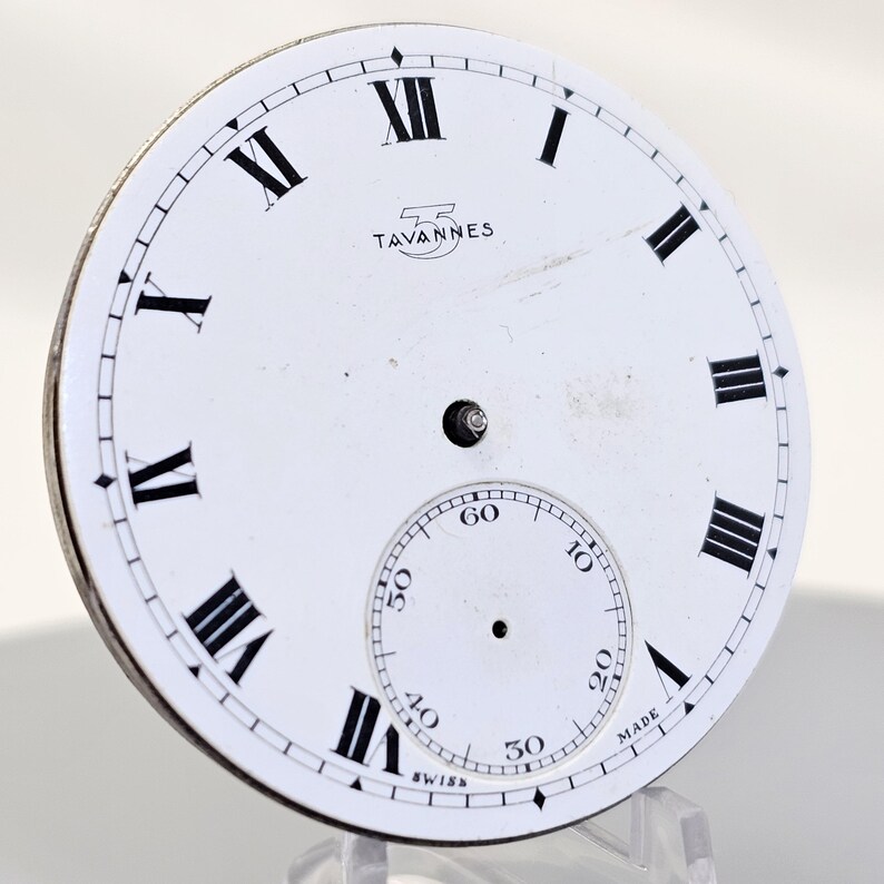 May include: A white watch face with black Roman numerals and a smaller seconds dial. The brand name "TAVANNES" is printed above the center. The word "SWISS" is printed at the bottom.