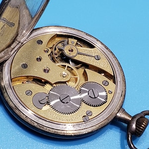 May include: A silver pocket watch with a gold-toned face and intricate gears visible through the open case. The watch has a black hand and a white dial with black markings.