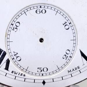 May include: Close-up of a white watch face with black numerals marking the hours and minutes. The watch face has a small hole in the center and the words "SWISS" and "MADE" are visible.